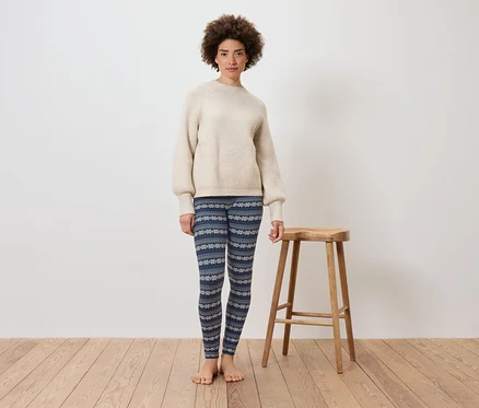 Eine Frau steht vor einer weißen Wand neben einem Holzhocker. Sie trägt einen cremefarbenen Pullover und blaue Leggings mit Muster.