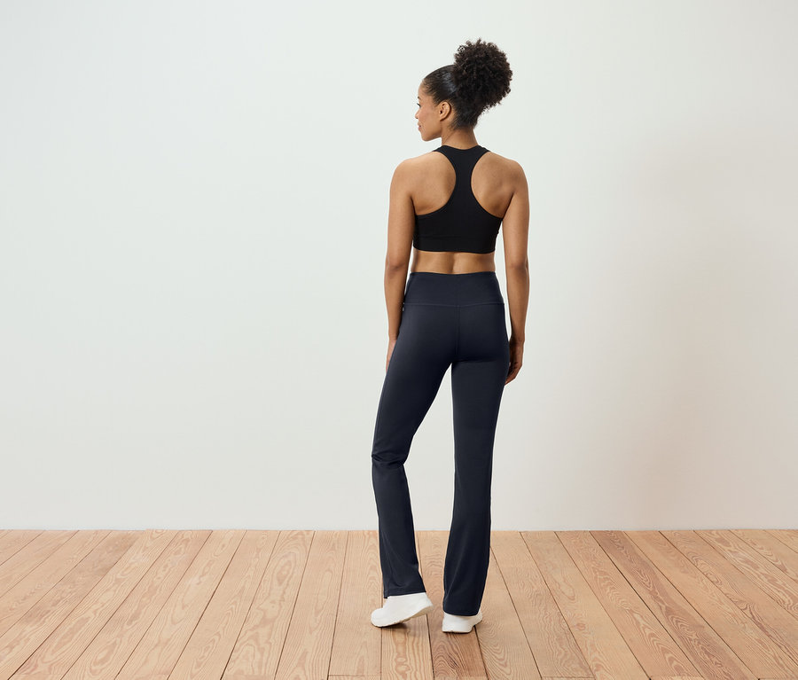 Rückansicht einer Frau mit hochgesteckten Haaren, die ein schwarzes Sportoberteil und eine dunkelblaue Hose trägt und auf einem Holzboden steht.