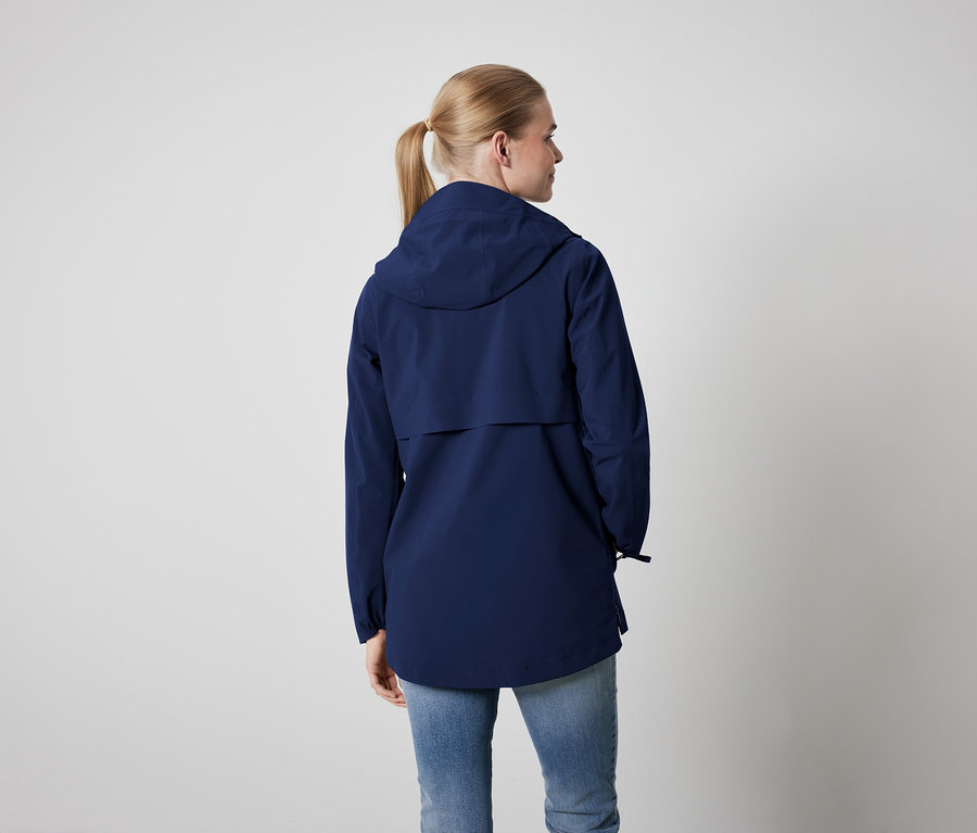Rückansicht einer Frau mit blonden, zu einem Pferdeschwanz zusammengebundenen Haaren, die eine blaue Jacke mit Kapuze und blaue Jeans trägt.