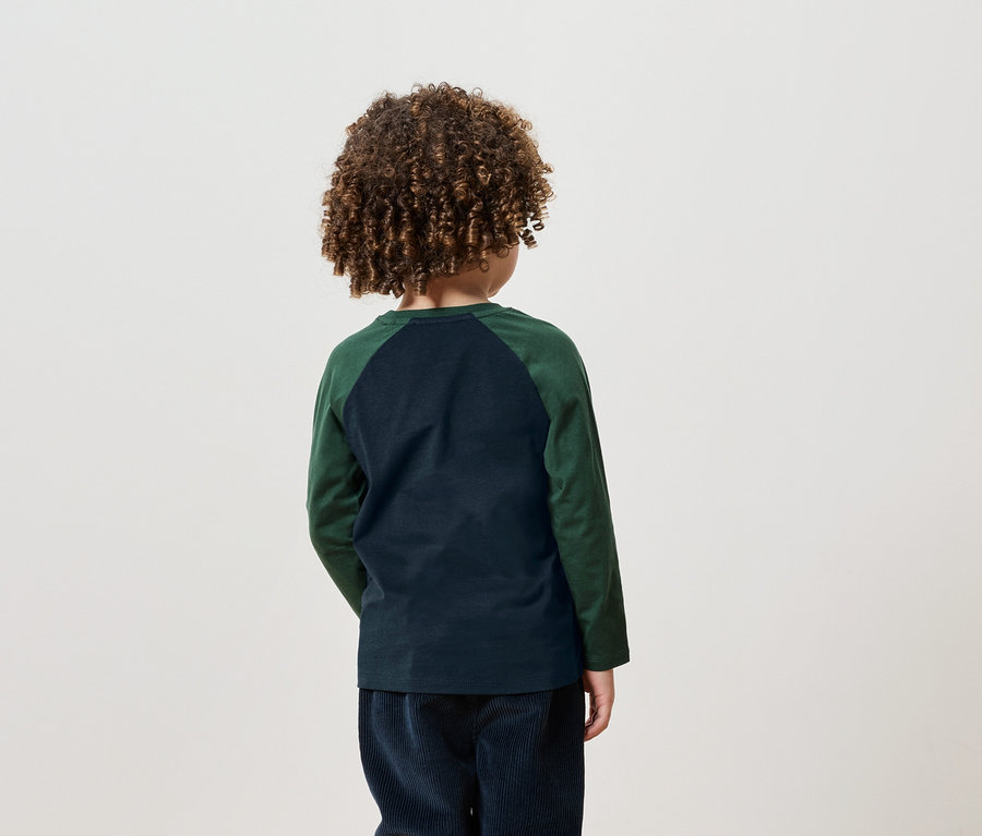 Junge mit lockigem Haar steht mit dem Rücken zur Kamera, trägt ein grünes Langarmshirt und marineblaue Cordhosen.