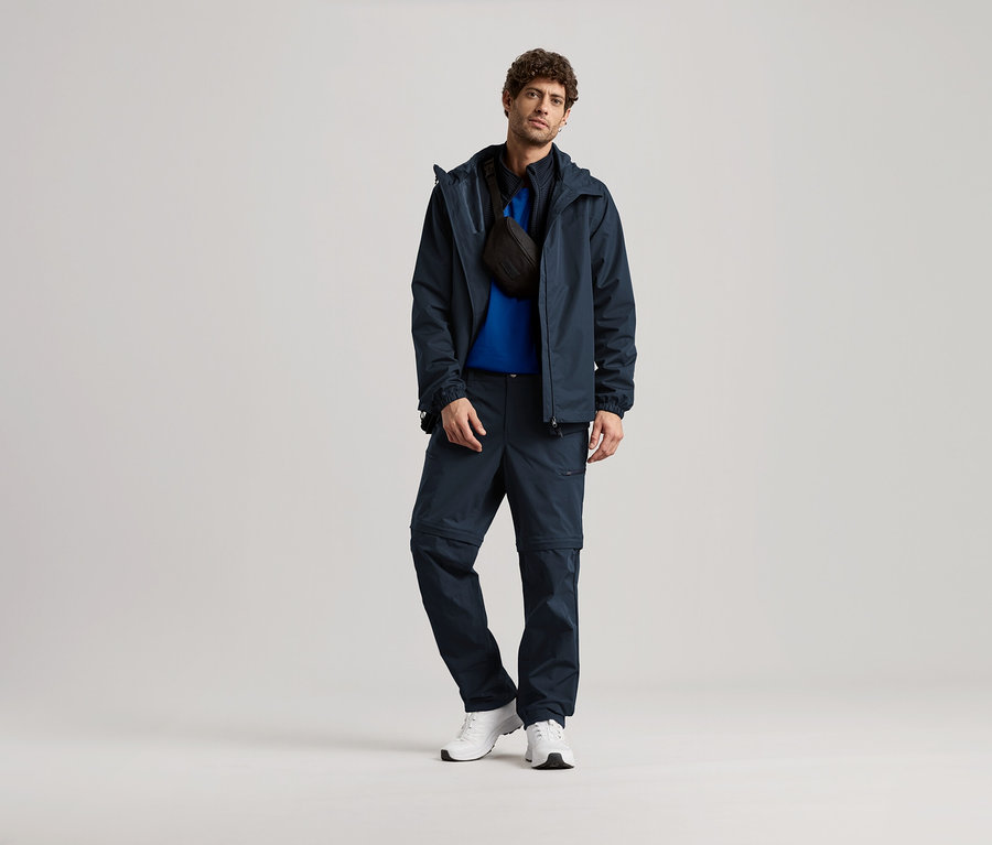 Ein Mann steht in marineblauer Jacke, Hose und weißen Turnschuhen und trägt eine schwarze Umhängetasche.