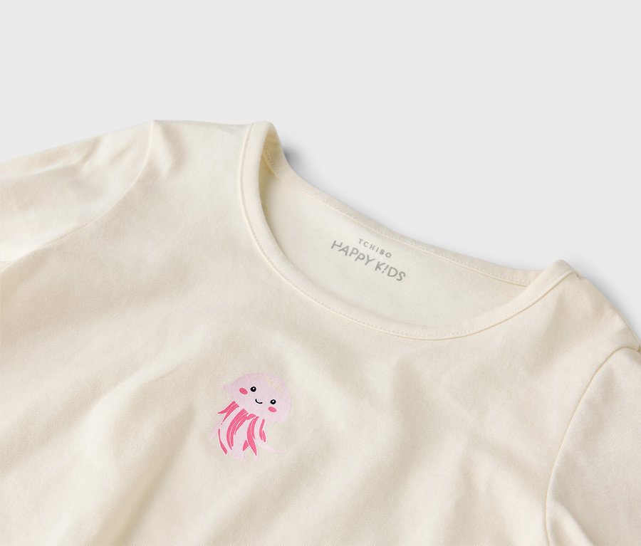 Nahaufnahme eines cremefarbenen T-Shirts mit einem rosafarbenen Quallen-Aufdruck und dem Aufdruck "Tchibo Happy Kids" im Nackenbereich.