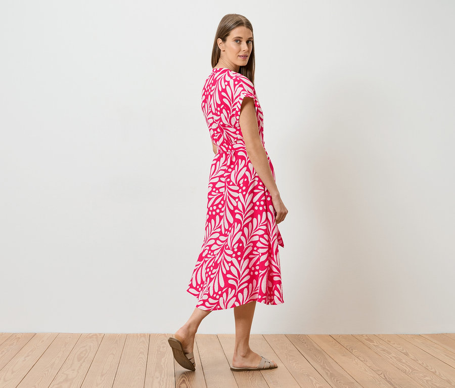Frau in einem pinkfarbenen Kleid mit weißem Blattmuster, die über einen Holzboden geht und über die Schulter blickt.