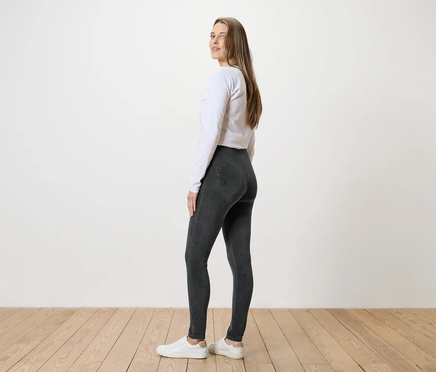 Frau mit langem Haar, weißem Oberteil, grauer Leggings und weißen Turnschuhen steht seitlich vor einer weißen Wand.