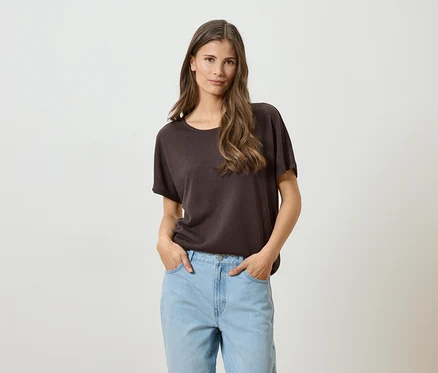 Frau mit langen braunen Haaren, braunem T-Shirt und hellblauen Jeans mit Händen in den Taschen vor einem weißen Hintergrund.