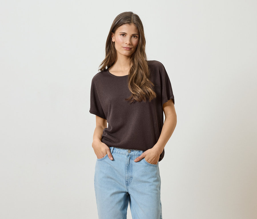 Frau mit langen braunen Haaren, braunem T-Shirt und hellblauen Jeans mit Händen in den Taschen vor einem weißen Hintergrund.