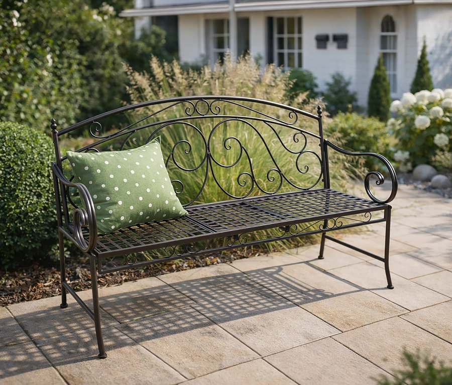Eine schwarze Metallbank mit einem grünen Kissen mit weißen Punkten steht auf einer Terrasse vor einem Haus.