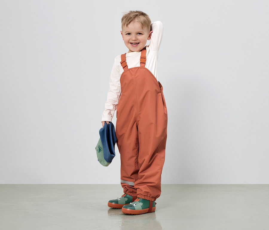 Ein lächelnder Junge mit hellbraunen Haaren trägt eine orangefarbene Latzhose, ein helles Hemd und grüne Gummistiefel mit Tiermotiven.