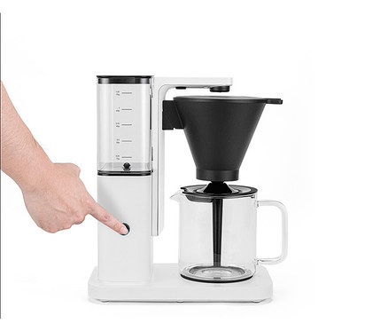 Hand drückt Knopf an weißer Kaffeemaschine mit Glaskanne und schwarzem Filter.