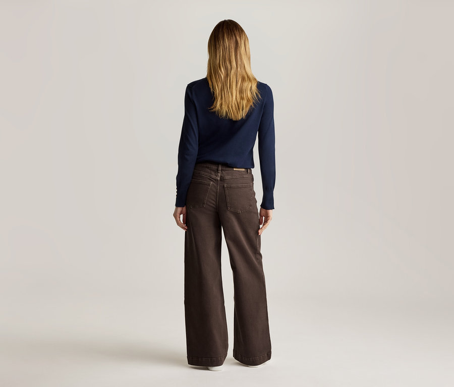 Von hinten ist eine Frau mit langen, blonden Haaren zu sehen, die einen marineblauen Pullover und eine braune, weite Hose trägt.