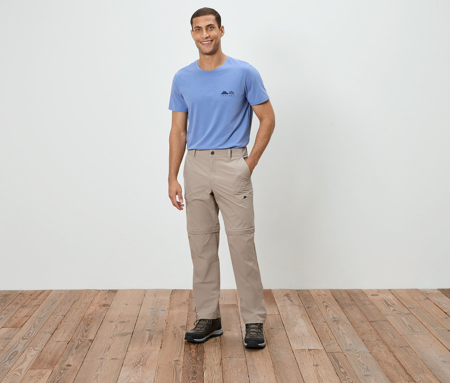 Ein Mann steht und trägt ein blaues T-Shirt, eine beige Hose und braune Schuhe.