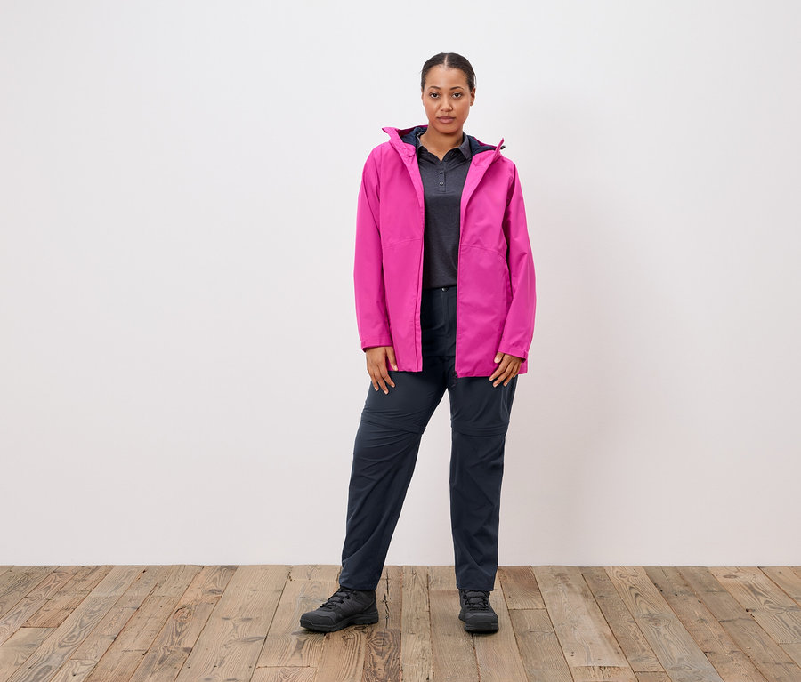 Frau steht vor einer weißen Wand und trägt eine pinke Jacke, ein graues Poloshirt und dunkelblaue Hosen.