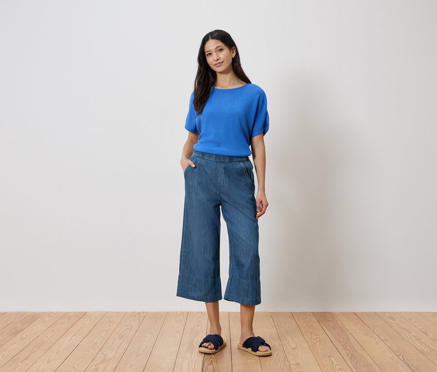 Frau steht mit Hand in der Hosentasche, trägt blaues Oberteil, Jeans-Culottes und Sandalen vor einer weißen Wand.