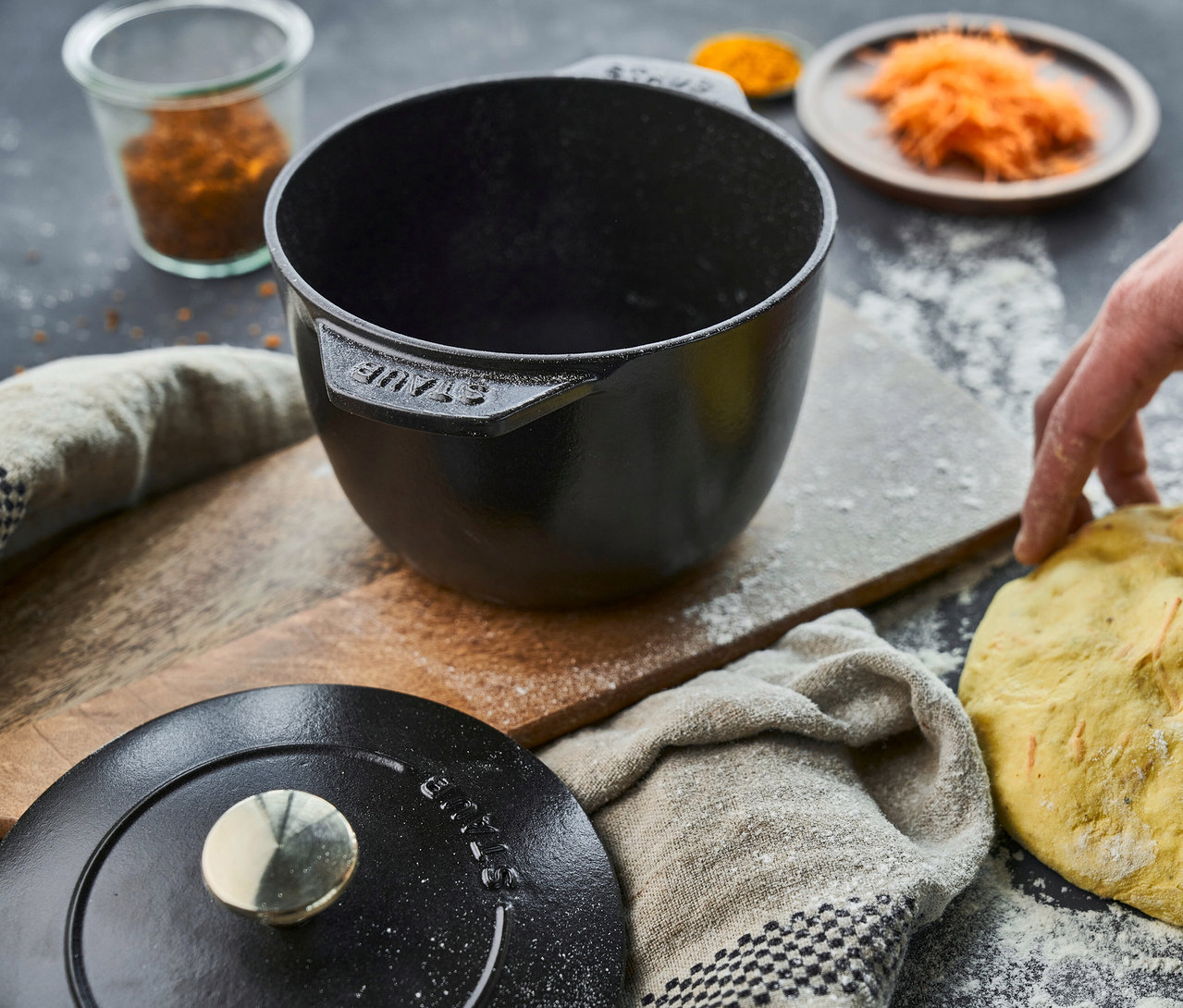 Eine schwarze, runde STAUB Reis-Cocotte (16 cm) steht auf einem Holztisch. Daneben liegen Teig, Gewürze und der Cocotte-Deckel.