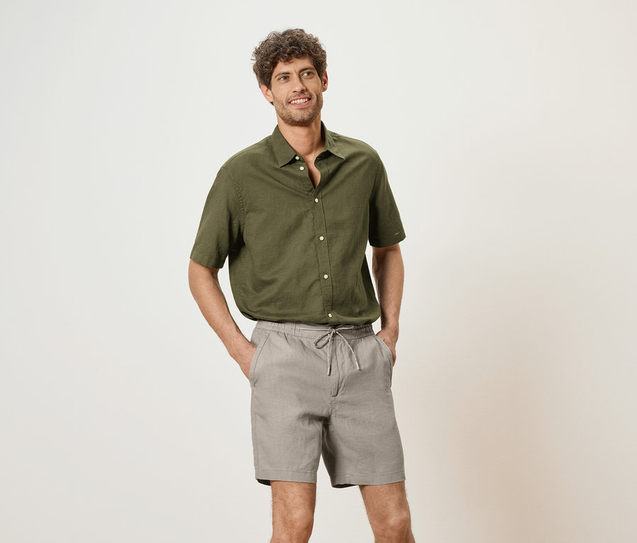 Mann mit olivgrünem Hemd und grauen Shorts, die Hände in den Hosentaschen, vor einem neutralen Hintergrund.