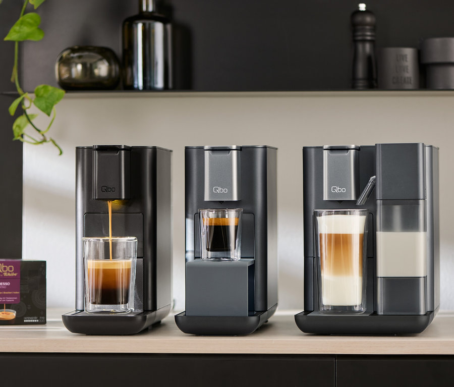 Drei Qbo Kaffeemaschinen stehen auf einer Theke, darunter Qbo MILK MASTER Gentle Grey, ICONIC Dark Chrome und ESSENTIAL Dark Stone. Espresso wird zubereitet.