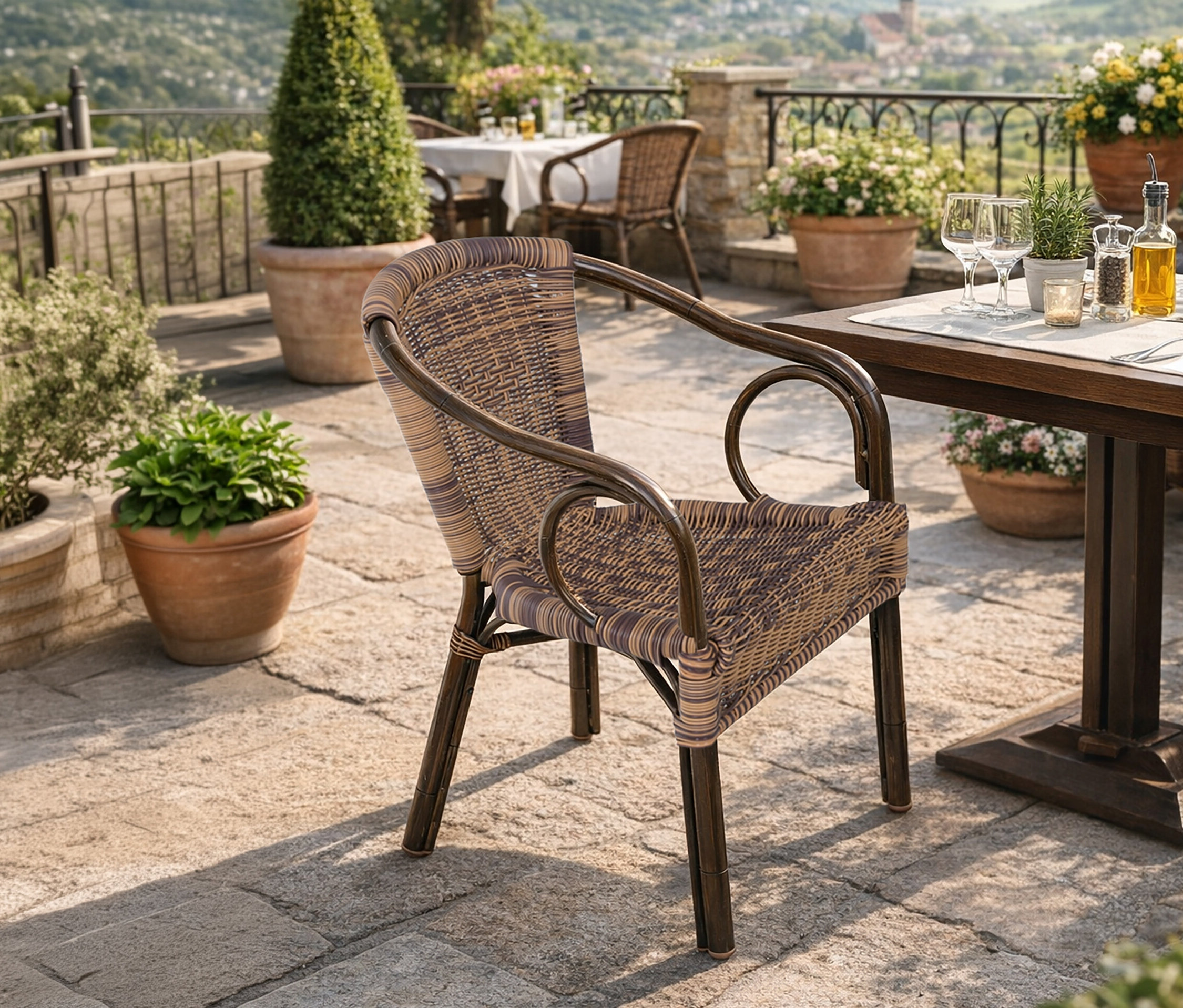 Ein brauner Rattanstuhl steht auf einer Terrasse mit Tischen, Stühlen und Topfpflanzen mit Blick auf eine Landschaft.