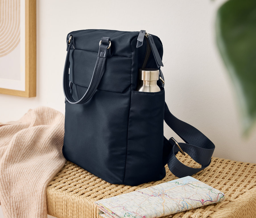 Eine dunkelblaue Rucksacktasche steht auf einer geflochtenen Bank. Eine silberne Wasserflasche steckt in einer Seitentasche der Tasche. Ein hellrosa Strickcardigan liegt auf der Bank, davor eine gefaltete Karte.