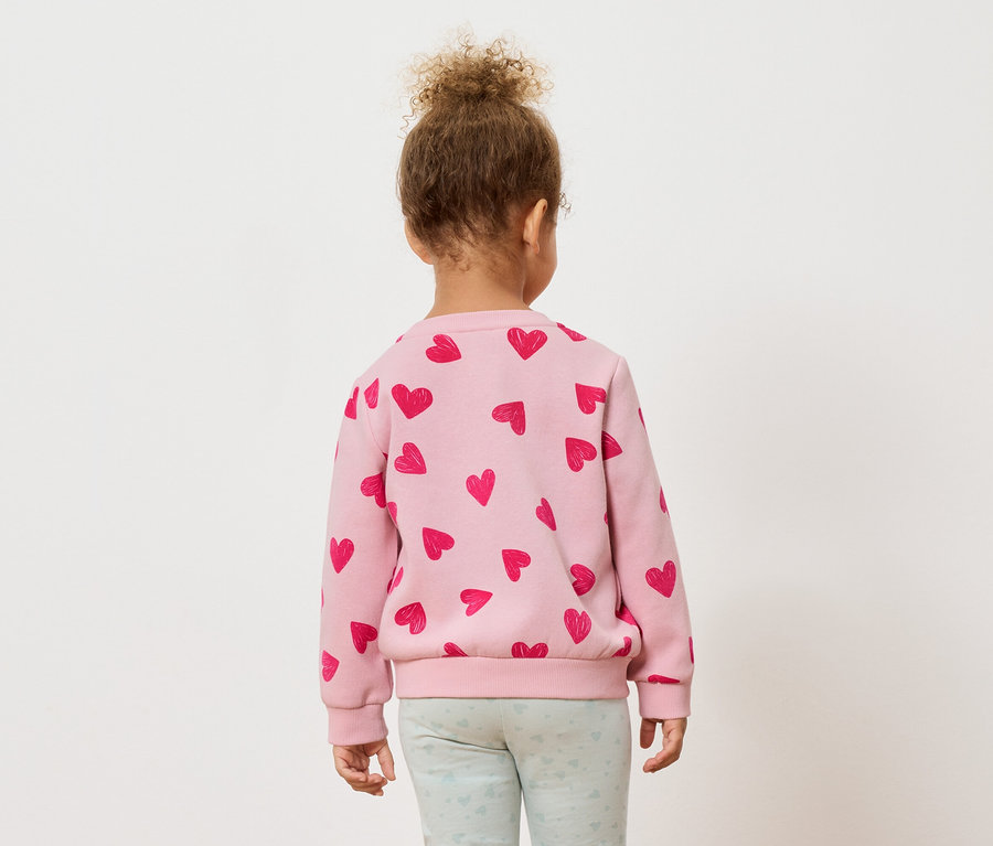 Rückansicht eines kleinen Kindes mit lockigem Haar, das einen rosa Pullover mit roten Herzen und hellblaue Leggings trägt.