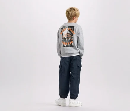 Ein Kind von hinten, trägt einen grauen Pullover mit "West Coast Surfing"-Aufdruck, marineblaue Cargohosen und weiße Turnschuhe.
