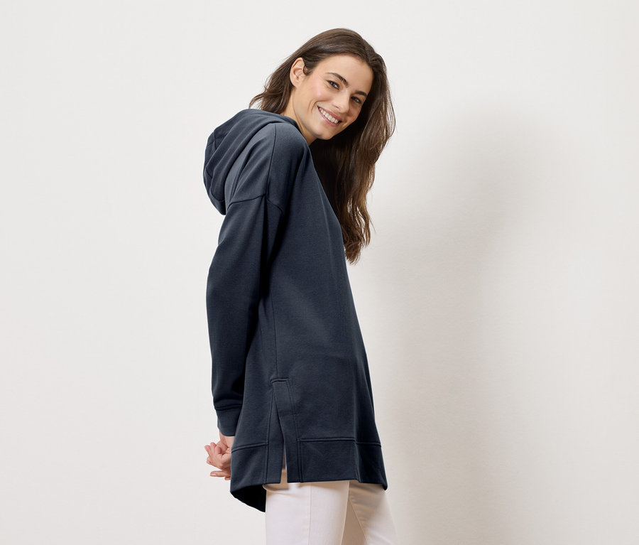 Frau trägt einen langen, marineblauen Hoodie und helle Hosen, lächelt in die Kamera.