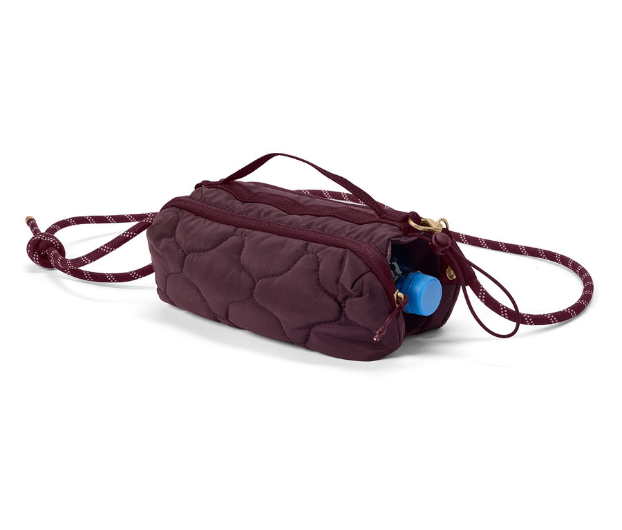 Eine auberginefarbene, gesteppte Zylindertasche mit Kordelzug und einer blauen Wasserflasche, die aus dem Reißverschluss herausschaut.