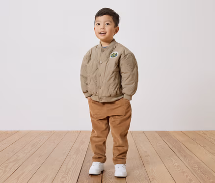 Ein lächelnder Junge mit beiger, gesteppter Jacke mit Aufnäher, Cordhose und weißen Schuhen steht auf einem Holzboden vor einer weißen Wand.