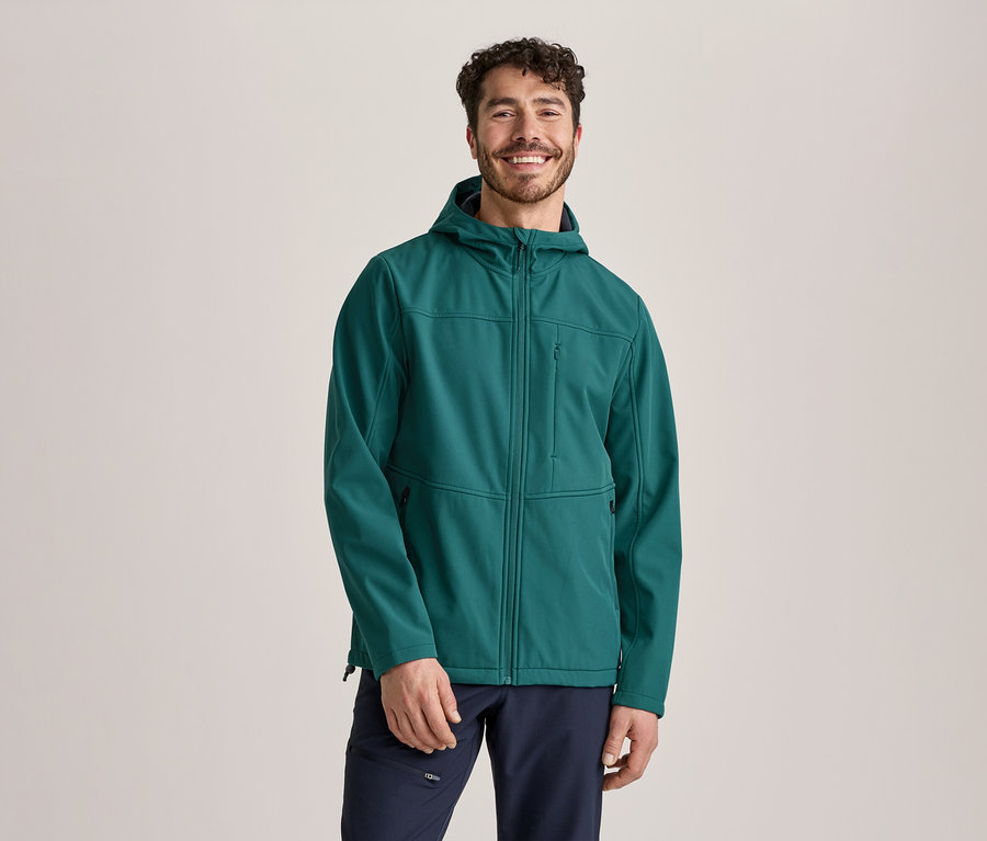 Ein lächelnder Mann mit lockigem Haar und Bart steht frontal da und trägt eine grüne Kapuzen-Softshelljacke und dunkelblaue Hose.