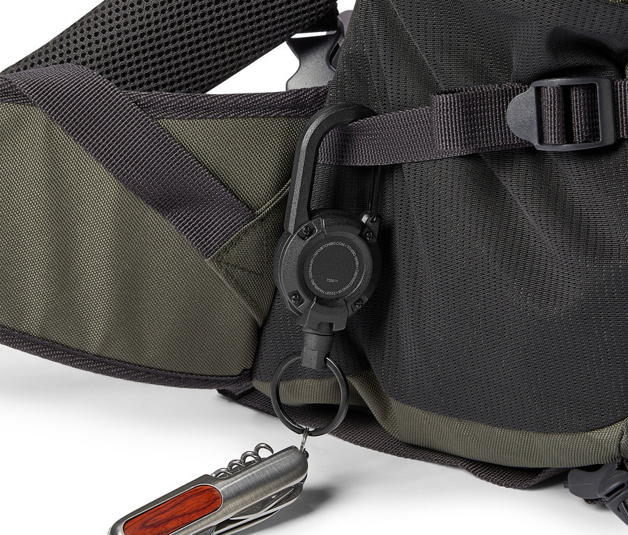 Nahaufnahme eines olivgrünen und schwarzen Rucksacks mit einem schwarzen Karabinerhaken, an dem ein Schlüsselanhänger und ein silbernes Taschenmesser mit Holzgriff befestigt sind.