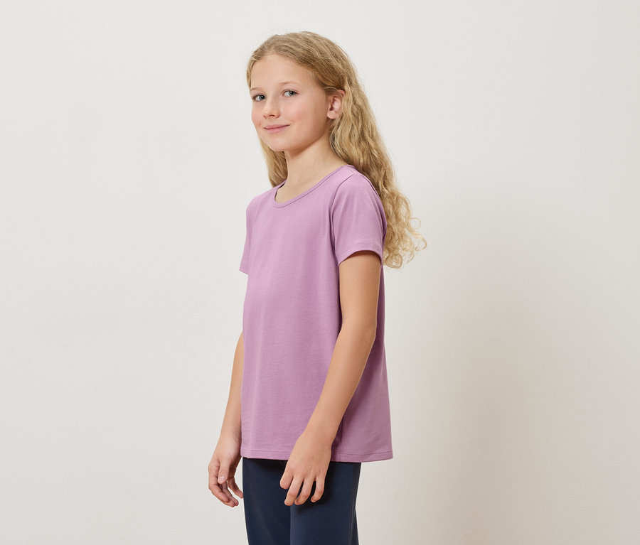 Mädchen mit langen blonden Haaren, das ein lila T-Shirt und eine dunkelblaue Hose trägt, steht vor einem weißen Hintergrund.