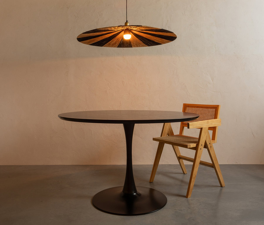 Ein runder schwarzer Tisch mit einem einzelnen Säulenfuß steht mittig, rechts daneben ein heller Holzstuhl mit geflochtener Sitzfläche und Rückenlehne. Über dem Tisch hängt eine flache, runde Pendelleuchte mit schwarz-naturfarbenen Streifen.