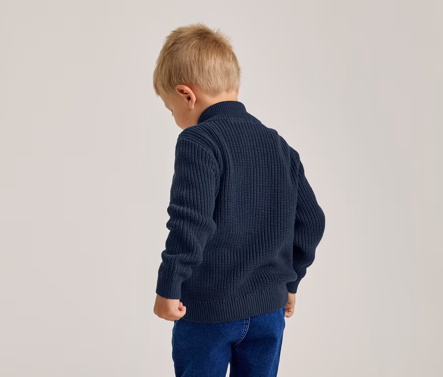 Junger Junge von hinten, trägt einen marineblauen Strickpullover und blaue Jeans, der Kopf ist leicht gesenkt.