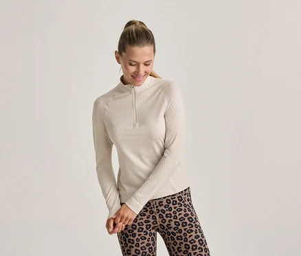 Lächelnde Frau mit Pferdeschwanz, die ein helles, geripptes Langarmshirt mit kurzem Reißverschluss und Leggings mit Leopardenmuster trägt.
