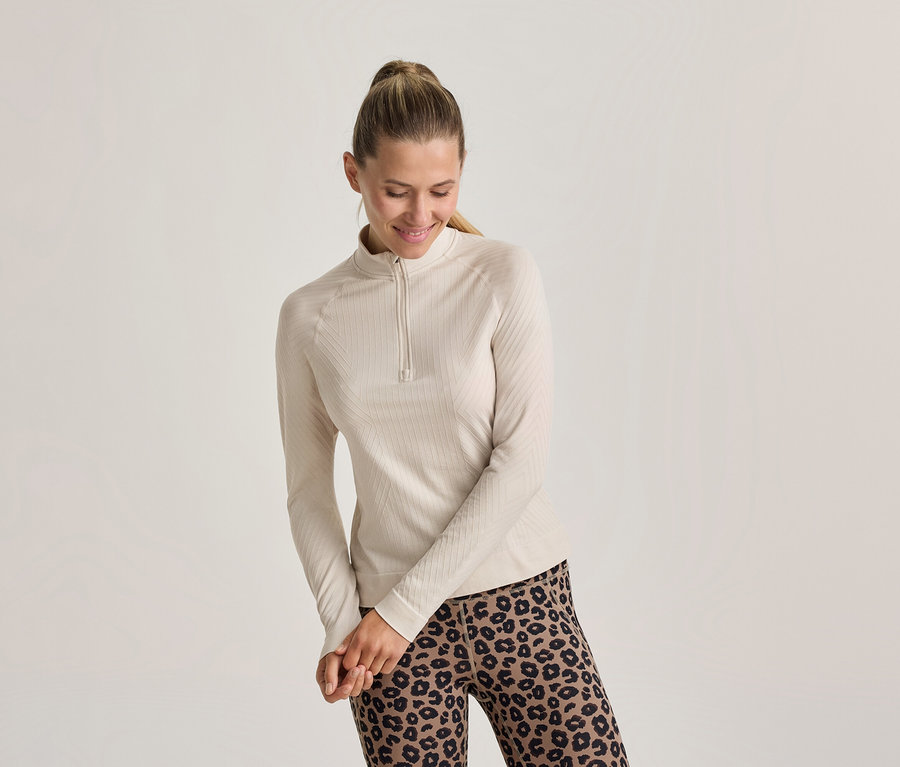 Lächelnde Frau mit Pferdeschwanz, die ein helles, geripptes Langarmshirt mit kurzem Reißverschluss und Leggings mit Leopardenmuster trägt.