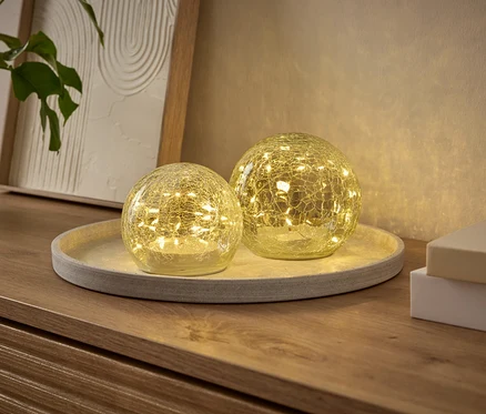 Zwei leuchtende, kugelförmige Dekoleuchten mit Craquelé-Effekt, gefüllt mit kleinen LEDs, stehen auf einem runden Tablett auf einer Holzkommode. Eine Pflanze und ein gerahmtes Bild sind im Hintergrund.
