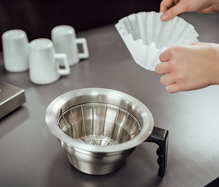 Hände legen einen Papierfilter in einen Metallfilter einer Kaffeemaschine. Im Hintergrund befinden sich drei weiße Tassen und eine silberne Basis einer Kaffeemaschine. Zusehen ist eine Profi-Filterkaffeemaschine »Tchibo Businessline«, ohne Kanne.
