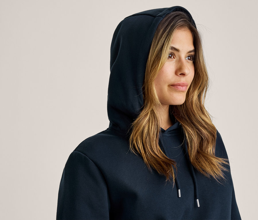Eine Frau mit hochgezogener Kapuze eines dunkelblauen Hoodies blickt nach rechts. Ihr langes, gewelltes braunes Haar fällt unter der Kapuze hervor.