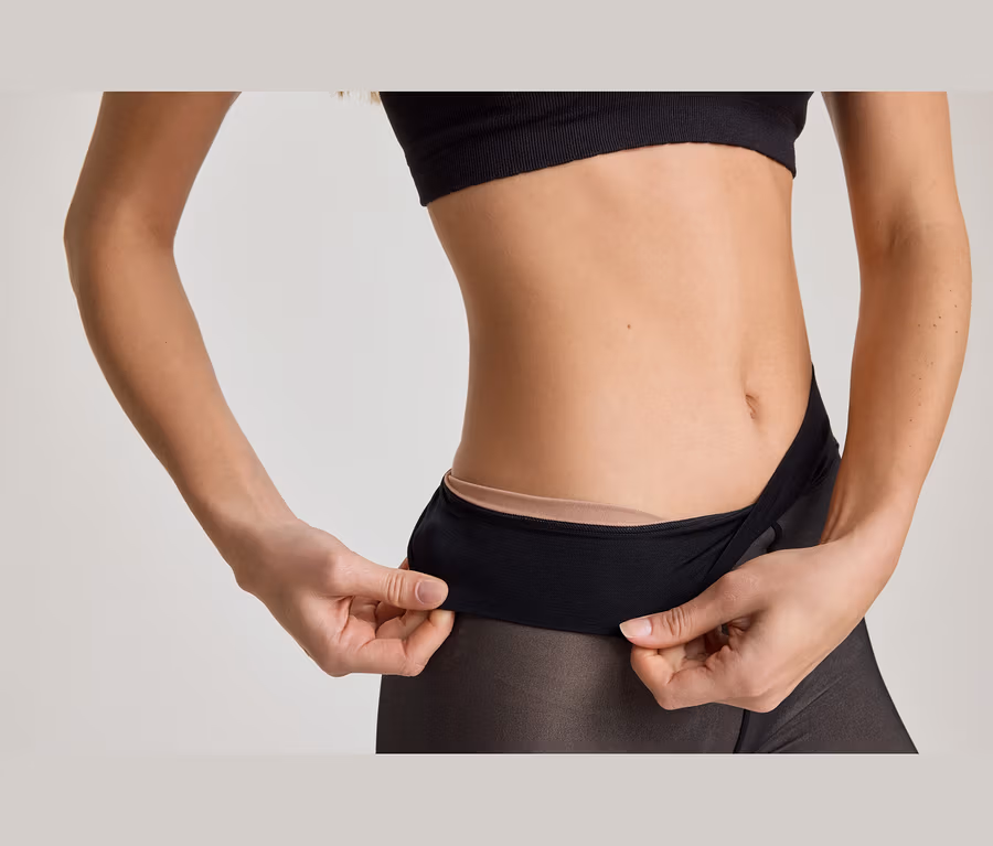 Oberkörper einer Person in schwarzem Sport-BH und transparenter schwarzer Strumpfhose, Hände ziehen am Bund, um hautfarbene Unterwäsche zu zeigen.