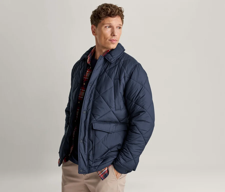 Mann mit kurzen lockigen Haaren trägt eine offene, marineblaue Steppjacke über einem rot-blauen Karoshirt und hellen Hosen, blickt nach rechts.