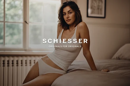 Eine Frau sitzt auf einem Bett, trägt ein weißes Tanktop und einen weißen Slip und blickt in die Kamera. Im Hintergrund sind ein Fenster und ein Heizkörper zu sehen.