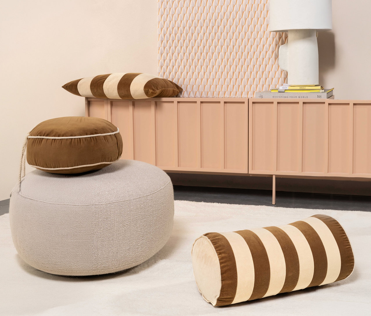Ein braunes Essenza Samt-Dekokissen »Bell« liegt auf einem beigen Pouf vor einem pfirsichfarbenen Schrank. Zwei weitere braun-beige gestreifte Samtkissen sind auf dem Schrank und dem weißen Teppich platziert.