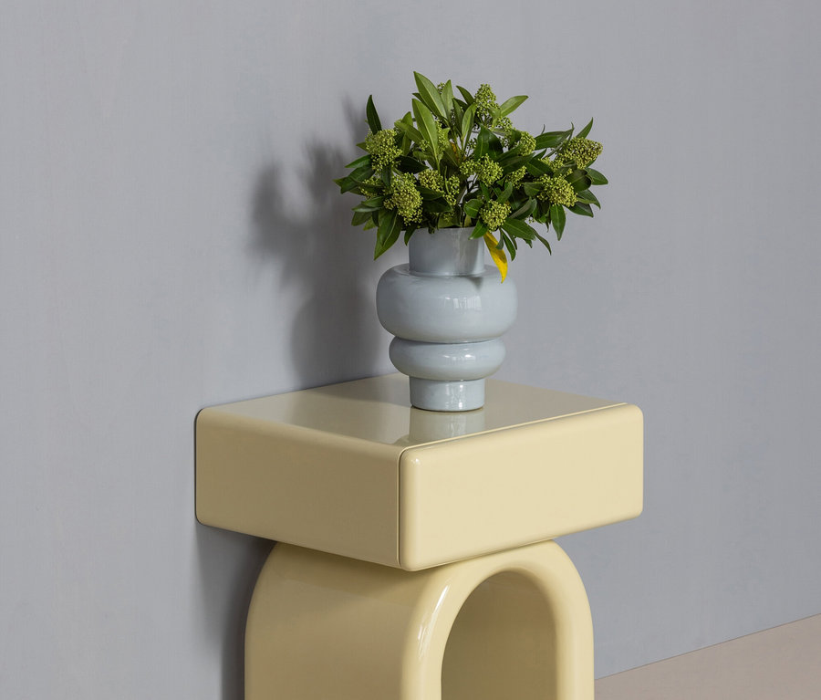 Ein hellgelber Beistelltisch mit gewölbter Basis und einer rechteckigen Platte steht vor einer hellgrauen Wand. Darauf steht eine hellblaue, bauchige Vase mit grünem Laub und kleinen Beeren.