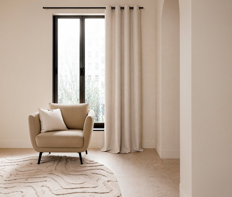 Ein beiger Sessel mit einem weißen Kissen steht vor einem Fenster. Rechts hängt ein beiger SCHÖNER WOHNEN-Kollektion Verdunkelungsvorhang »OPACO« an einer schwarzen Stange. Auf dem Boden liegt ein runder, beiger Teppich mit Wellenmuster.