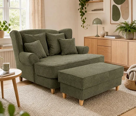 Ein gemütliches Wohnzimmer mit einer grünen Récamiere mit mehreren Kissen und einem passenden Hocker auf einem hellen Teppich. Links steht ein Holztischchen mit einer Tasse, rechts ein Holz-Sideboard mit einer grünen Lampe, Pflanzen und abstrakter Kunst.