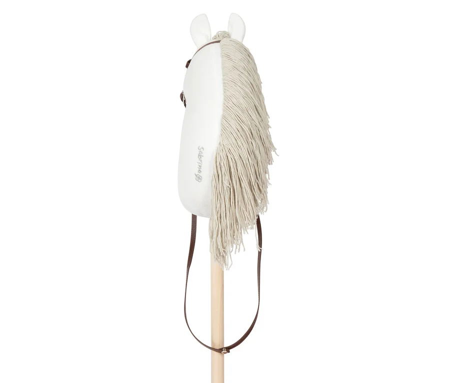 Weißes Steckenpferd mit hellbeiger Garnmähne, braunem Zaumzeug und Holzstab. Ein "Sabrina"-Logo ist auf dem weißen Plüschkopf aufgestickt. Ein brauner Riemen hängt am Zaumzeug herunter.