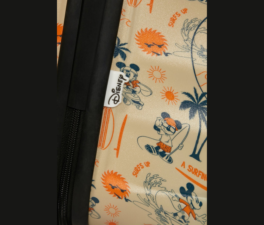 Nahaufnahme eines hellen Koffers mit einem sommerlichen Mickey-Mouse-Muster. Das Muster zeigt Mickey beim Surfen, Skateboardfahren und Entspannen, zusammen mit Palmen, Sonnen und Surfbrettern. Ein schwarzer Reißverschluss und ein weißes Disney-Etikett sind links sichtbar.