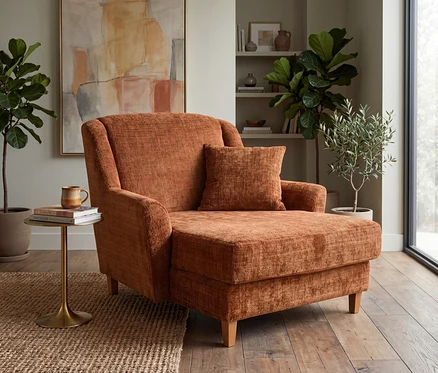 Ein rostfarbener Chaiselongue-Sessel mit passendem Kissen steht auf einem Jute-Teppich und Holzboden. Ein goldener Beistelltisch daneben trägt eine Tasse und Bücher. Im Hintergrund hängen ein abstraktes Gemälde und ein Bücherregal mit Deko ist neben einem Fenster zu sehen.