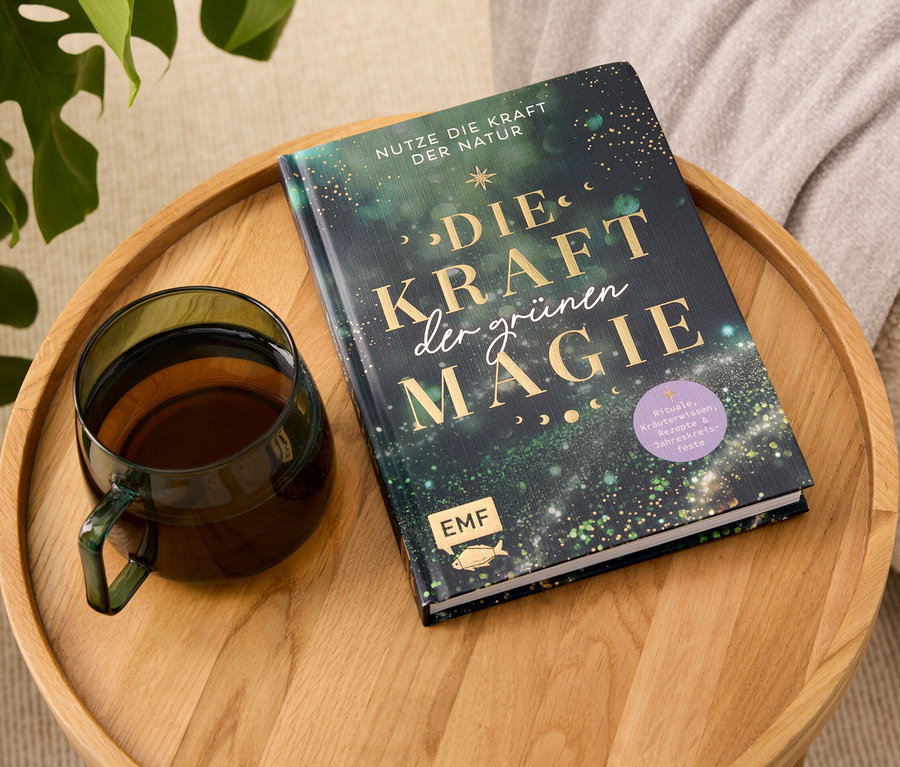 Auf einem Holztablett liegen ein Buch mit dem Titel "Die Kraft der grünen Magie" und eine gläserne Tasse mit Getränk.