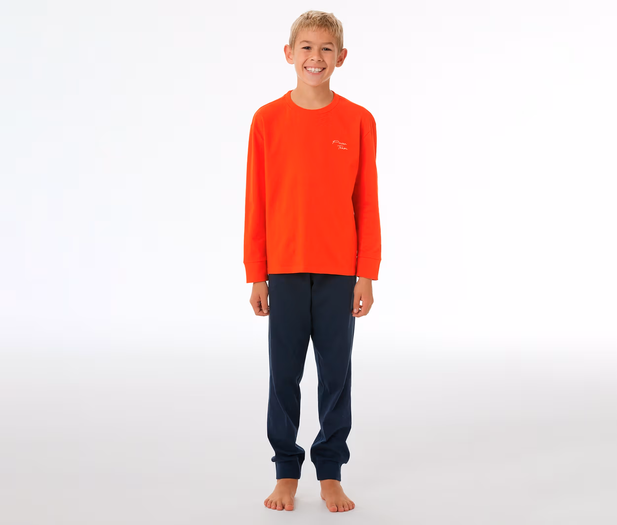 Lächelnder Junge in einem orangefarbenen SCHIESSER Kinder-Pyjama mit Bündchen und dunkelblauer Hose, barfuß stehend.