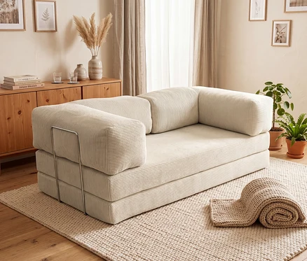 Ein hellbeiges Modulsofa mit Cordstruktur und Armlehnenstützen aus Metall steht auf einem strukturierten Teppich in einem gemütlichen Wohnzimmer. Dahinter befindet sich ein Holzsideboard mit Büchern, Gläsern und Pampasgras. Topfpflanzen stehen auf dem Holzboden, gerahmte botanische Drucke hängen an der Wand neben einem Fenster mit hellen Vorhängen. Eine zusammengerollte Strickdecke liegt auf dem Teppich.